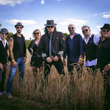 EIne Band, die in einem herbstlichen Acker steht: vorne der Bandleader, der aussieht wie Udo Lindenberg mit Hut und Sonnenbrille