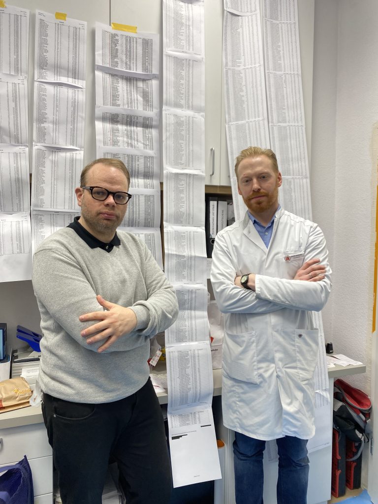 MdB Helge Lindh und Inhaber David Kudlek stehen in der Wuppertaler Klingelholl-Apotheke vor den seitenlangen Listen vergriffener Medikamente