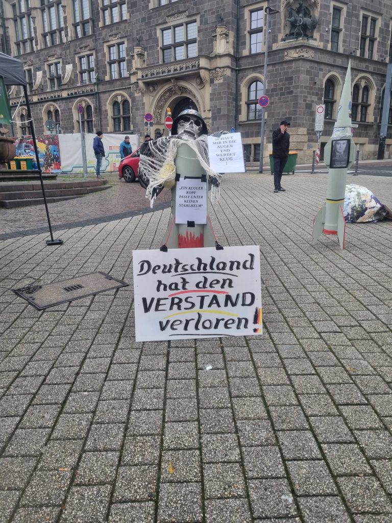 Nachbau einer Rakete, darauf ein Totenkopf mit Stahlhelm, mit dem Text: „Schaut mir in die Augen und werdet klug!“ Auf der Rakete steht ein Zitat von Albert Einstein: „Ein kluger Kopf passt unter keinen Stahlhelm“. Vor der Rakete steht ein Schild: „Deutschland hat den Verstand verloren!“