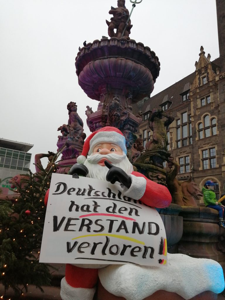 Foto: Ein Weihnachtsmann vor dem Brunnen am Neumarkt hält ein Schild: „Deutschland hat den Verstand verloren!“