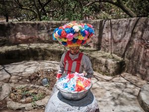 Eine südafrikanische Performerin trägt einen großen, festlichen Blumenhut und schaut in eine Schale voller Blumen.