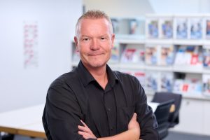 Porträt von Thorsten Schumacher, Geschäftsführer Operativ der Agentur für Arbeit Solingen-Wuppertal im BiZ. Er trägt ein schwarzes Oberhemd und verschränkt seine Arme vor der Brust.