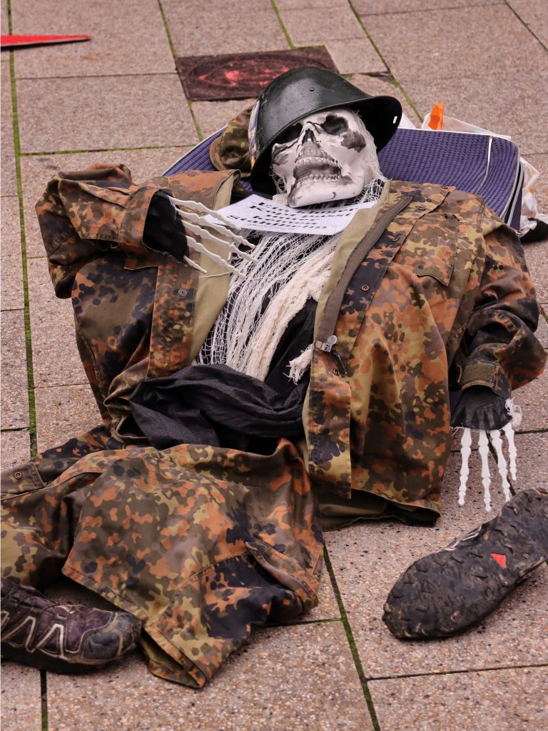 Foto: Ein Skelett mit Stahlhelm und einem Plakat: „Ein kluger Kopf passt unter keinen Stahlhelm“