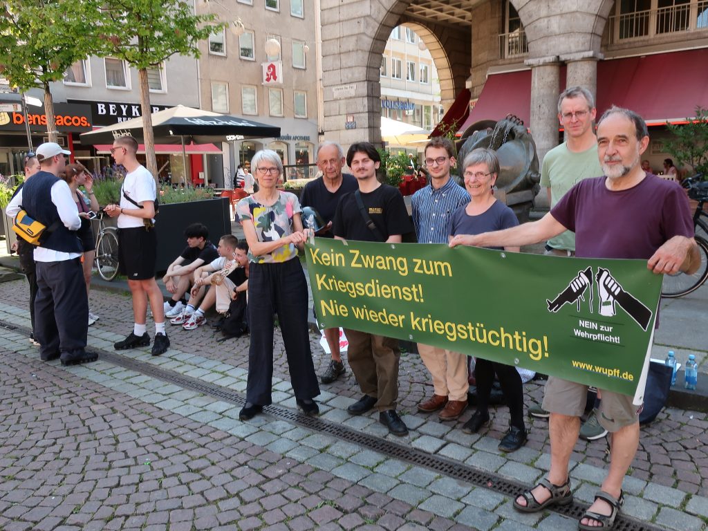 Foto mit dem Infostand, Demonstrierenden und Plakaten, auf einem sind Fotos von Soldaten, Panzern und einem Schützengraben im Krieg, dazu der Text: "Romantik auf dem Schlachtfeld"