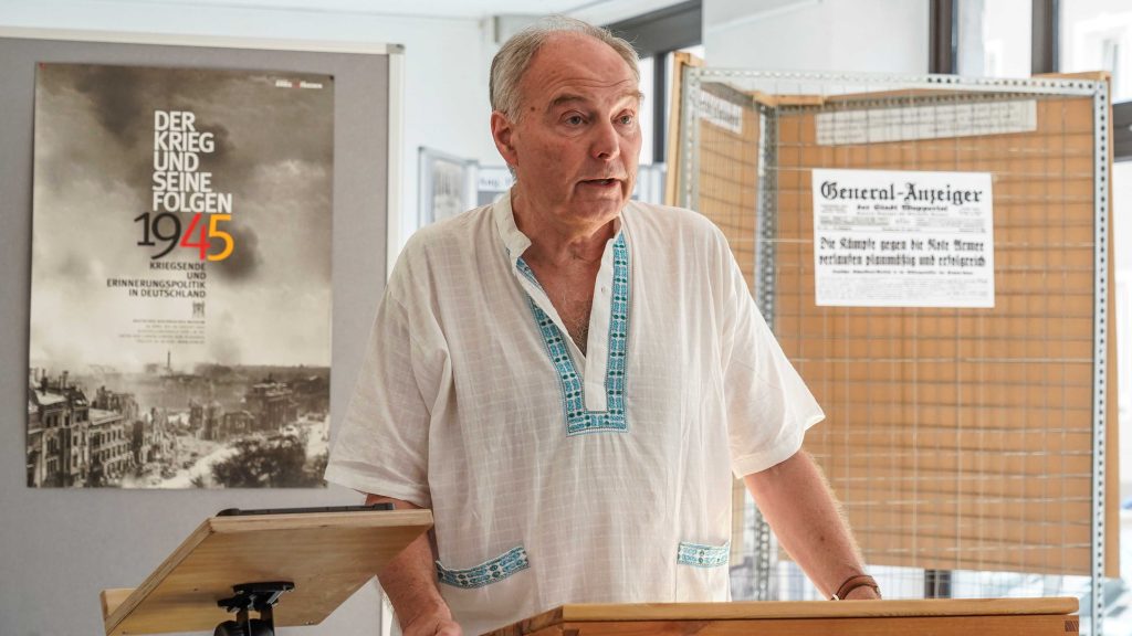 Foto: Reiner Rhefus am Rednerpult vor einem Plakat: „Der Krieg und seine Folgen 1945 – Kriegsende und Erinnerungspolitik in Deutschland“ mit einem Bild einer zerstörten Stadt